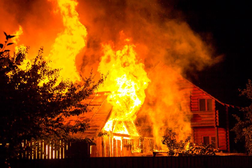 burning wooden house at night bright orange flames and dense smoke from under the tiled roof on dark sky trees silhouettes and residential neighbor cottage disaster and danger concept